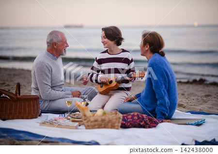 Grandparents enjoying beach picnic with teenage granddaughter playing ukulele by sea. Grandparents enjoying beach picnic with teenage granddaughter playing ukulele by sea. 142438000