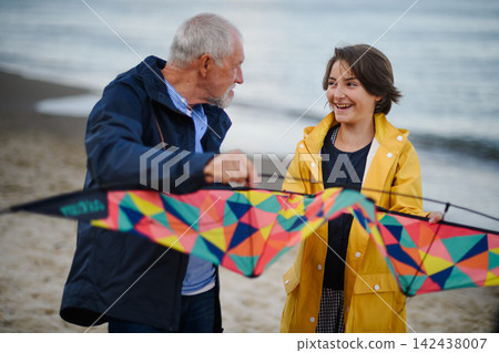 Senior man and his preteen granddaughter preparing kite for flying on sandy beach. Senior man and his preteen granddaughter preparing kite for flying on sandy beach. 142438007