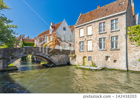 Bruges town in Belgium, Europe. Historic European Canal Scene with Ancient Stone Bridge Arching Over Tranquil Water, Flanked by Traditional Brick Buildings Under Clear Blue Sky 142438203