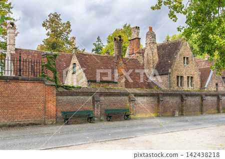 Bruges town in Belgium, Europe. Historic European Brick Buildings with Ornate Chimneys and Terracotta Roofs Behind a Long Brick Wall and Benches 142438218