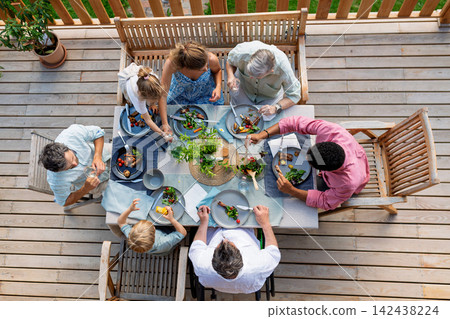 Above shot of family eating at barbecue party dinner on patio, little girl eating roasted corn and enjoying it. Above shot of family eating at barbecue party dinner on patio, little girl eating roasted corn and enjoying it. 142438224