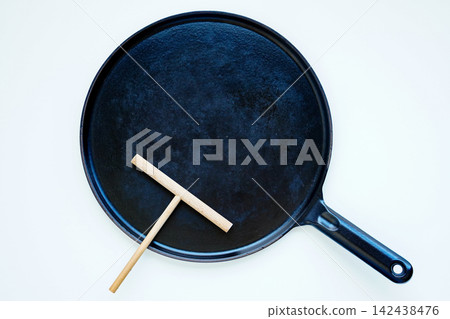 A black crepe pan and a crepe spreader, photographed from directly above against a white background. A black crepe pan and a crepe spreader, photographed from directly above against a white background. 142438476