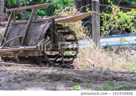 Mud covered walking tiller metal cage wheel resting beside rural rice field after soil preparation agricultural work Mud covered walking tiller metal cage wheel resting beside rural rice field after soil preparation agricultural work 142439988