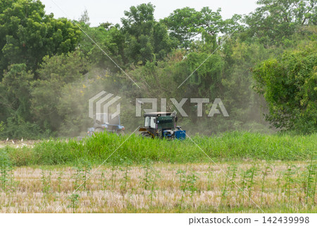 Two farm tractors working across rural field surrounded by green vegetation trees dust rising behind agricultural machinery during land cultivation Two farm tractors working across rural field surrounded by green vegetation trees dust rising behind agricultural machinery during land cultivation 142439998