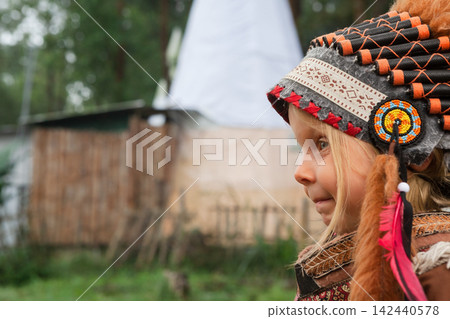 Portrait of young girl wearing a traditional Indian feathered costume. The image was taken outdoors in a forest like setting, evoking a cultural or educational experience for children. Portrait of young girl wearing a traditional Indian feathered costume. The image was taken outdoors in a forest like setting, evoking a cultural or educational experience for children. 142440578