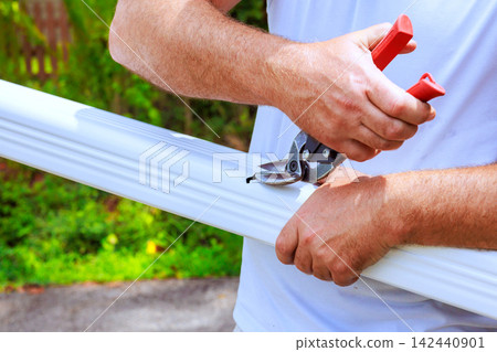 Worker uses sharp shears to cut piece of gutter metal pipe in garden area 142440901