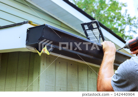 Contractor attaches gutter to edge of building while using drill with house with green siding. Contractor attaches gutter to edge of building while using drill with house with green siding. 142440904