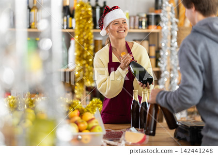 Woman in Santa hat offers customer to buy champagne or sparkling wine for Christmas celebration Woman in Santa hat offers customer to buy champagne or sparkling wine for Christmas celebration 142441248