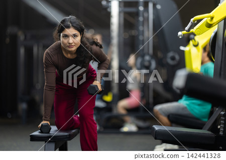 Latina woman doing dumbbell rows near bench in hall Latina woman doing dumbbell rows near bench in hall 142441328