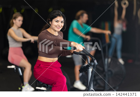 Positive Latina girl doing air bike exercises in well-equipped gym Positive Latina girl doing air bike exercises in well-equipped gym 142441371