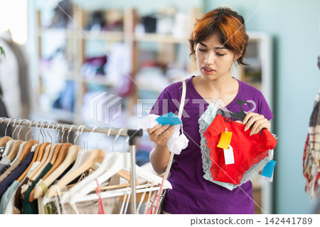 Girl looking at pair of lace-trimmed panties in clothing shop Girl looking at pair of lace-trimmed panties in clothing shop 142441789