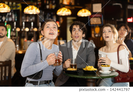 Attentive men and women watching sports match on TV, drinking beer and eating crisps, spending time together in bar Attentive men and women watching sports match on TV, drinking beer and eating crisps, spending time together in bar 142441797