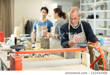 Elderly man in pottery workshop works with machine clay press roller Elderly man in pottery workshop works with machine clay press roller 142442006