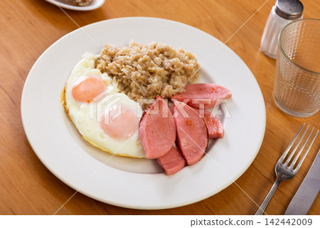 Sausages with fried egg and oatmeal on white plate. Breakfast option with eggs and porridge next to cutlery and glass of tea Sausages with fried egg and oatmeal on white plate. Breakfast option with eggs and porridge next to cutlery and glass of tea 142442009