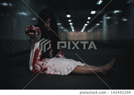Halloween theme featuring scary woman sitting on floor wearing bloody white gown inside dark corridor reaching hand forward during horror night celebration event activity Halloween theme featuring scary woman sitting on floor wearing bloody white gown inside dark corridor reaching hand forward during horror night celebration event activity 142442294