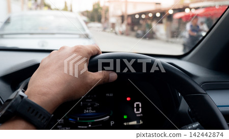 Driver is perspective holding a steering wheel inside a car. Stopped in heavy urban traffic during daytime. Parked behind the sedan in front of the road. Driver is perspective holding a steering wheel inside a car. Stopped in heavy urban traffic during daytime. Parked behind the sedan in front of the road. 142442874