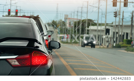 Rear side of black car stop and quere on asphalt road. Car turn on break light. Side road with electric pole and town include trees. on opposite with alone of car driving through. environment at days. Rear side of black car stop and quere on asphalt road. Car turn on break light. Side road with electric pole and town include trees. on opposite with alone of car driving through. environment at days. 142442883