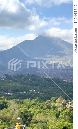 Mountain silhouette above volcanic plain under cloudy blue sky with foreground trees and village, with mountain, volcano, plain, cloud detail, showing sky, peak, silhouette, ridge context Mountain silhouette above volcanic plain under cloudy blue sky with foreground trees and village, with mountain, volcano, plain, cloud detail, showing sky, peak, silhouette, ridge context 142445242