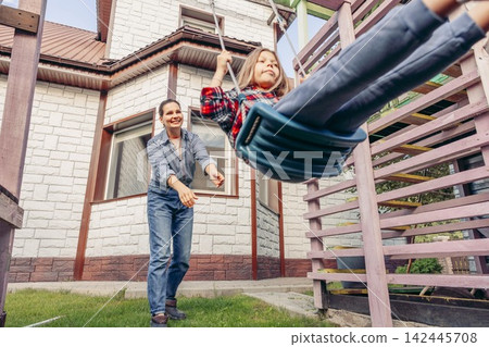 mother holding daughter on rope swing looking at camera in backyard garden, country house, happy family bonding moment outdoors, childhood joy, safety scene, family care, real estate mother holding daughter on rope swing looking at camera in backyard garden, country house, happy family bonding moment outdoors, childhood joy, safety scene, family care, real estate 142445708