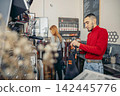 A young Caucasian man with short hair prepares coffee at a cafe. A young woman with long red hair stands behind him. The cafe has a modern design and coffee equipment. 142445776