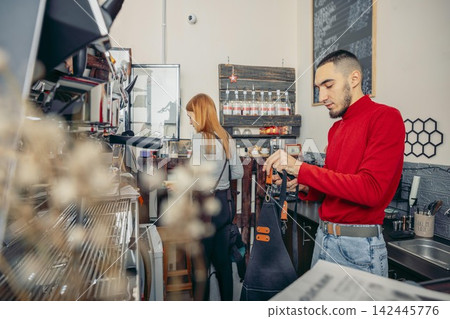 A young Caucasian man with short hair prepares coffee at a cafe. A young woman with long red hair stands behind him. The cafe has a modern design and coffee equipment. 142445776