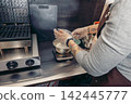 A barista prepares milk in a cafe. The person has tattoos and wears gloves while measuring ingredients on a scale. A waffle maker is visible in the background. 142445777