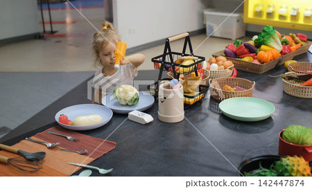 Little girl playing at an improvised kitchen with numerous toy ingredients and dishes preparing a meal from toy cabbage. Pretend cooking concept 142447874