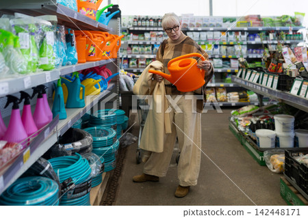 Senior woman shopping for gardening supplies, holding an orange watering can in a store aisle 142448171