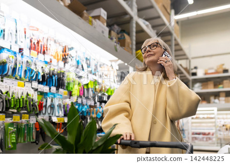 Mature woman smiling, engaging in a phone call, pushing a shopping cart in a garden center Mature woman smiling, engaging in a phone call, pushing a shopping cart in a garden center 142448225
