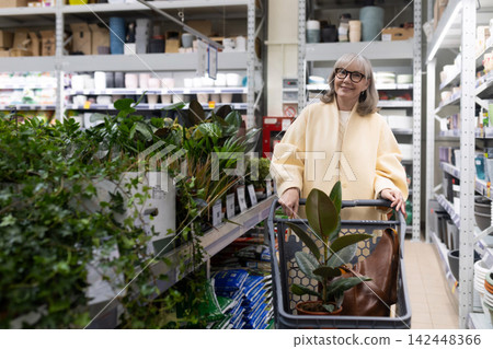 Mature woman smiling, pushing a shopping cart filled with a plant at a nursery Mature woman smiling, pushing a shopping cart filled with a plant at a nursery 142448366
