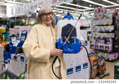 Senior woman shopping for gardening tools, happily holding a backpack sprayer in a hardware store Senior woman shopping for gardening tools, happily holding a backpack sprayer in a hardware store 142448384