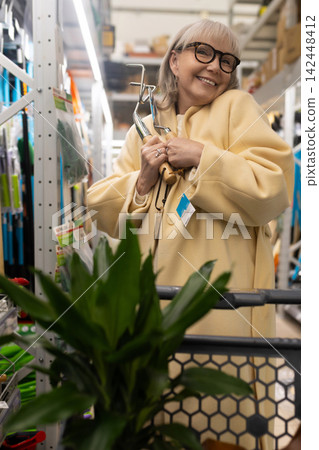 Senior woman smiling, holding gardening tools while shopping in a hardware store aisle Senior woman smiling, holding gardening tools while shopping in a hardware store aisle 142448412