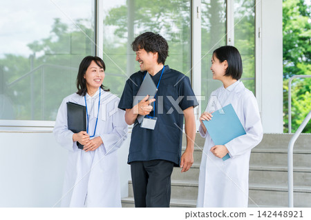Three medical staff members, some in white coats and others in scrubs, walk along, smiling and chatting. Three medical staff members, some in white coats and others in scrubs, walk along, smiling and chatting. 142448921