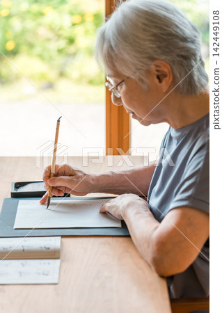 A senior woman sits at a desk practicing calligraphy with a brush. A senior woman sits at a desk practicing calligraphy with a brush. 142449108