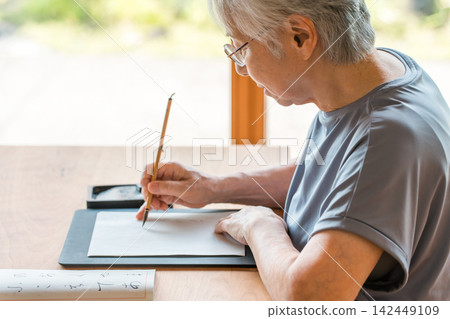 A senior woman sits at a desk practicing calligraphy with a brush. A senior woman sits at a desk practicing calligraphy with a brush. 142449109