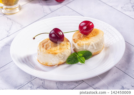 Two golden syrniki topped with fresh cherries and mint on white plate 142450009