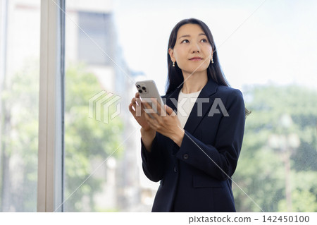 A young Japanese businesswoman is using her smartphone by a bright window. A young Japanese businesswoman is using her smartphone by a bright window. 142450100