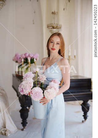 Woman in light blue dress holding peonies by piano Woman in light blue dress holding peonies by piano 142450223