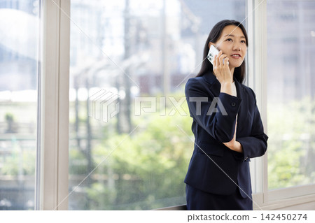 A young Japanese businesswoman talks on her smartphone by a bright window. A young Japanese businesswoman talks on her smartphone by a bright window. 142450274