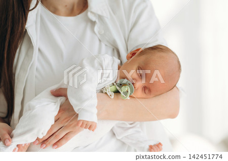 Mother holding sleeping infant in white outfit with pacifier, soft natural light illuminating the cozy indoor setting, showcasing a nurturing moment between parent and child Mother holding sleeping infant in white outfit with pacifier, soft natural light illuminating the cozy indoor setting, showcasing a nurturing moment between parent and child 142451774