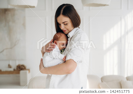 Young woman holding newborn baby in her arms, dressed in white shirt, standing in bright, modern living room with soft furnishings and natural light 142451775