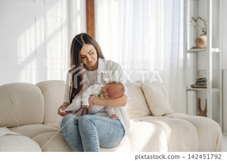 Young woman in casual attire holds a newborn baby while seated on a cozy sofa in a bright, modern living room with natural light streaming through the windows Young woman in casual attire holds a newborn baby while seated on a cozy sofa in a bright, modern living room with natural light streaming through the windows 142451792