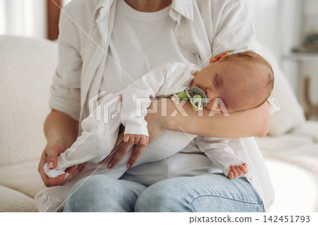Caucasian woman in white shirt holds sleeping baby in white outfit with pacifier while seated on a light-colored sofa in a bright indoor setting Caucasian woman in white shirt holds sleeping baby in white outfit with pacifier while seated on a light-colored sofa in a bright indoor setting 142451793