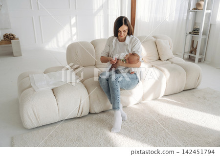 Woman sitting on a modern sofa in a bright room, gently holding and feeding an infant while surrounded by soft furnishings and natural light 142451794