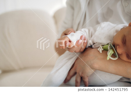 Close-up of a parent holding a sleeping baby in white clothing, with a pacifier attached, seated on a light-colored sofa in a softly lit room Close-up of a parent holding a sleeping baby in white clothing, with a pacifier attached, seated on a light-colored sofa in a softly lit room 142451795
