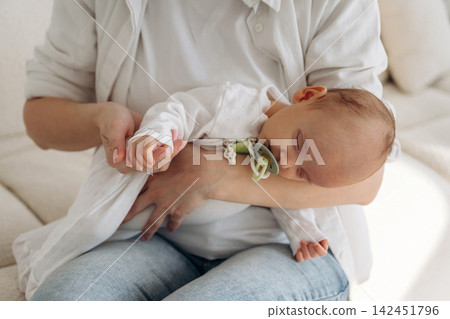 Caucasian woman in white shirt holds sleeping infant in her arms while seated on a light-colored sofa in a cozy indoor setting with soft lighting 142451796