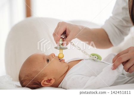 Infant lying on a soft surface while a caregiver gently places a pacifier in the baby's mouth, with a bright and airy background enhancing the scene Infant lying on a soft surface while a caregiver gently places a pacifier in the baby's mouth, with a bright and airy background enhancing the scene 142451811