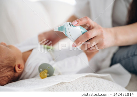 Caregiver using nasal aspirator on infant lying on soft blanket, pacifier nearby, with gentle lighting creating a calm atmosphere in a cozy indoor setting Caregiver using nasal aspirator on infant lying on soft blanket, pacifier nearby, with gentle lighting creating a calm atmosphere in a cozy indoor setting 142451814