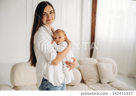 Young Caucasian mother holding her infant child in a bright, modern living room with soft furnishings and natural light streaming through sheer curtains Young Caucasian mother holding her infant child in a bright, modern living room with soft furnishings and natural light streaming through sheer curtains 142451837