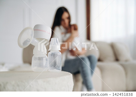 Breast pump and baby bottle on a table in a bright living room with a mother holding her infant in the background, soft natural light filtering through the window Breast pump and baby bottle on a table in a bright living room with a mother holding her infant in the background, soft natural light filtering through the window 142451838